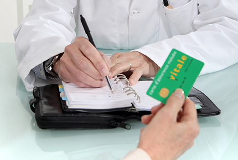 2 Photographie d'une main tenant une carte vitale de la sécurité sociale avec les mains d'un médecin écrivant dans un agenda dans le cadre du plafond mensuel de la sécurité sociale en 2021