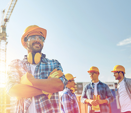 Un ouvrier en premier plan sourit et 3 ouvriers derrière lui discutent ensemble au milieu d'un chantier avec leurs casques, lunettes et gants de protections dans le cadre de la présentation des valeurs et de l'engagement de la complémentaire santé de la mutuelle MBTP